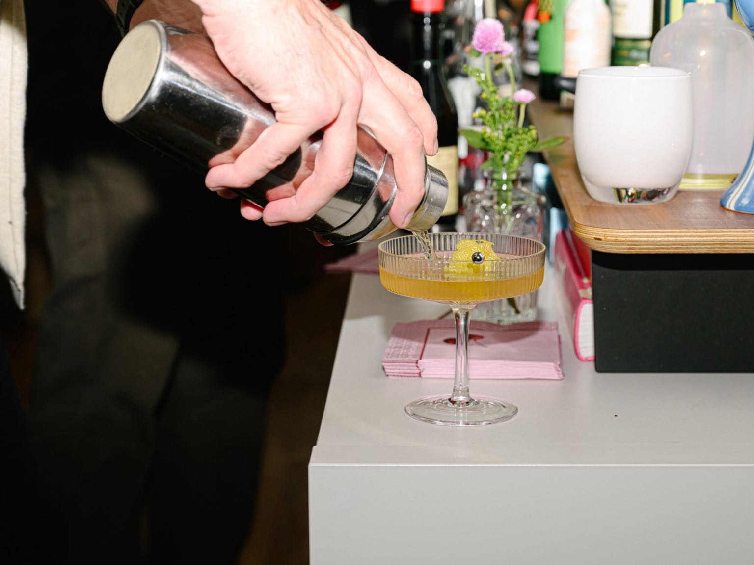 A cocktail being poured from a shaker into a coupe with a lemon twist